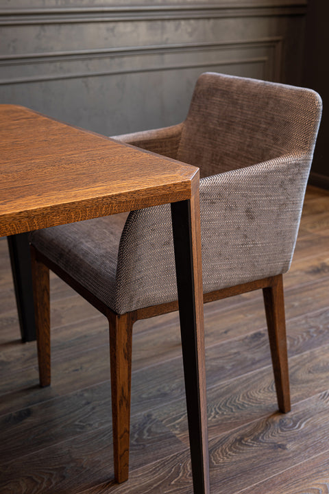 Fragment drewnianego stołu i szarego tapicerowanego krzesła przy ciemnej ścianie.